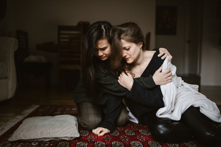 Two women sit closely together indoors, offering comfort and support to each other.