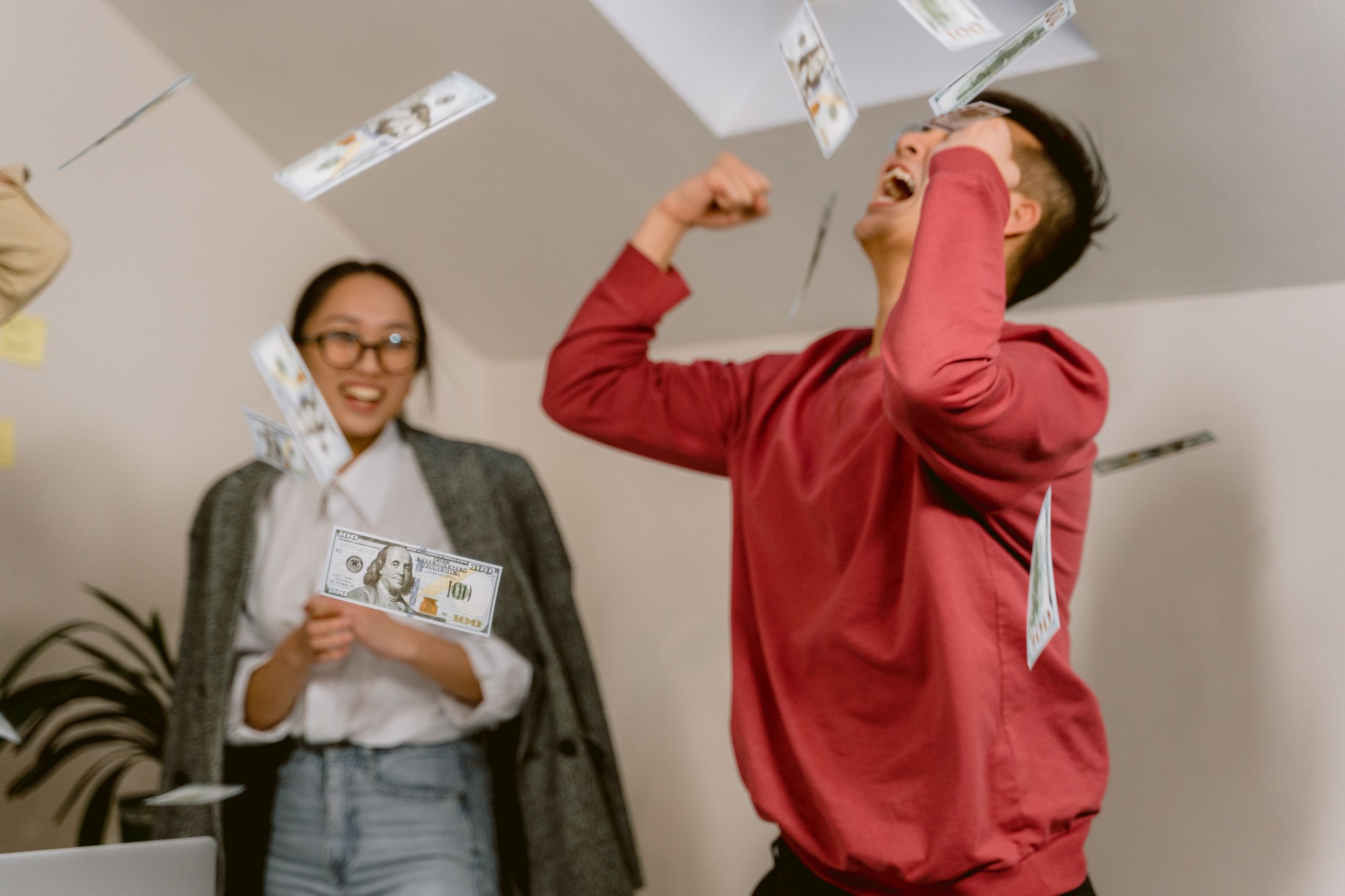 Two young adults in a room celebrating with falling dollar bills, expressing excitement and joy.