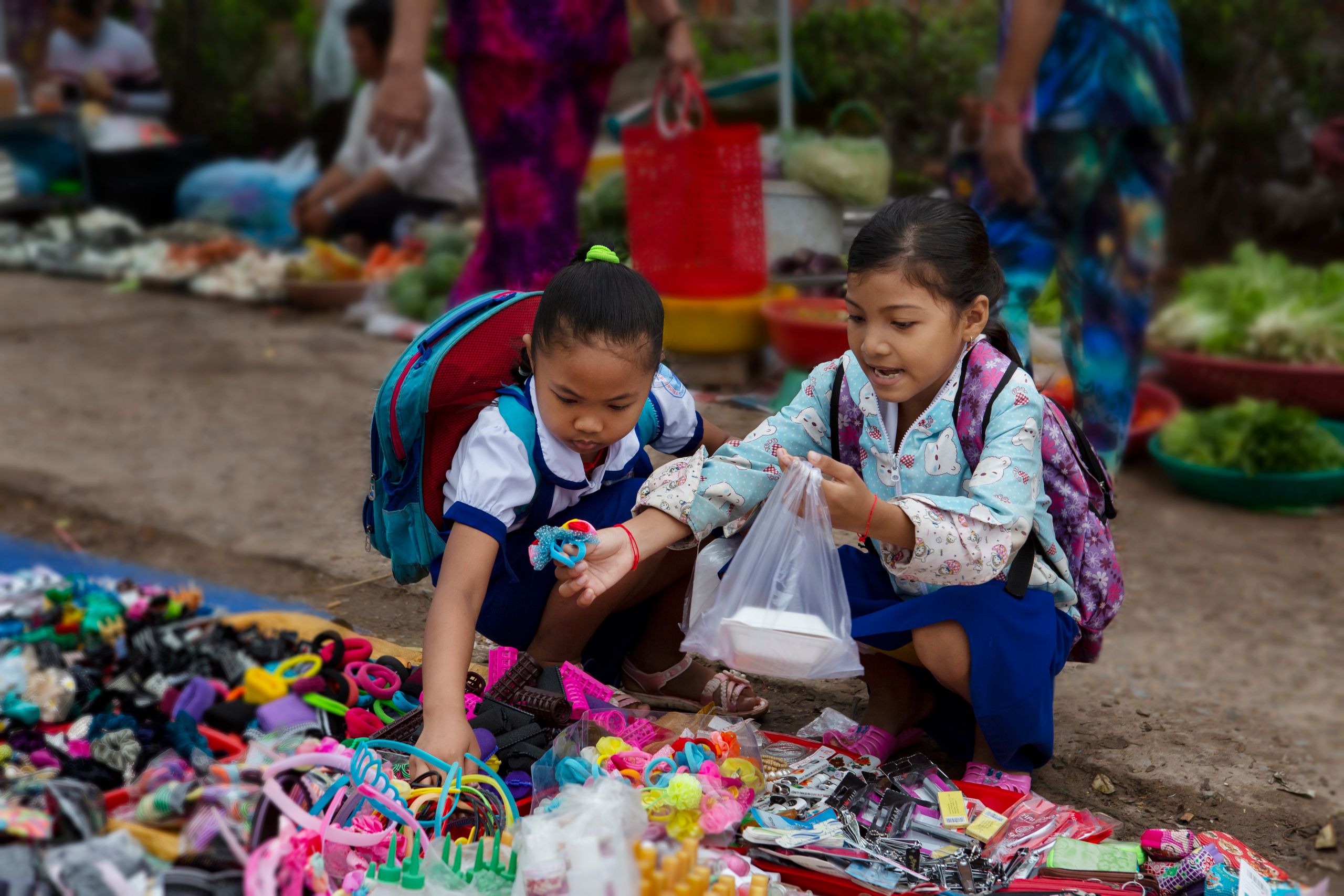 Two young schoolgirls explore and purchase items at a lively outdoor market, filled with vibrant colors.