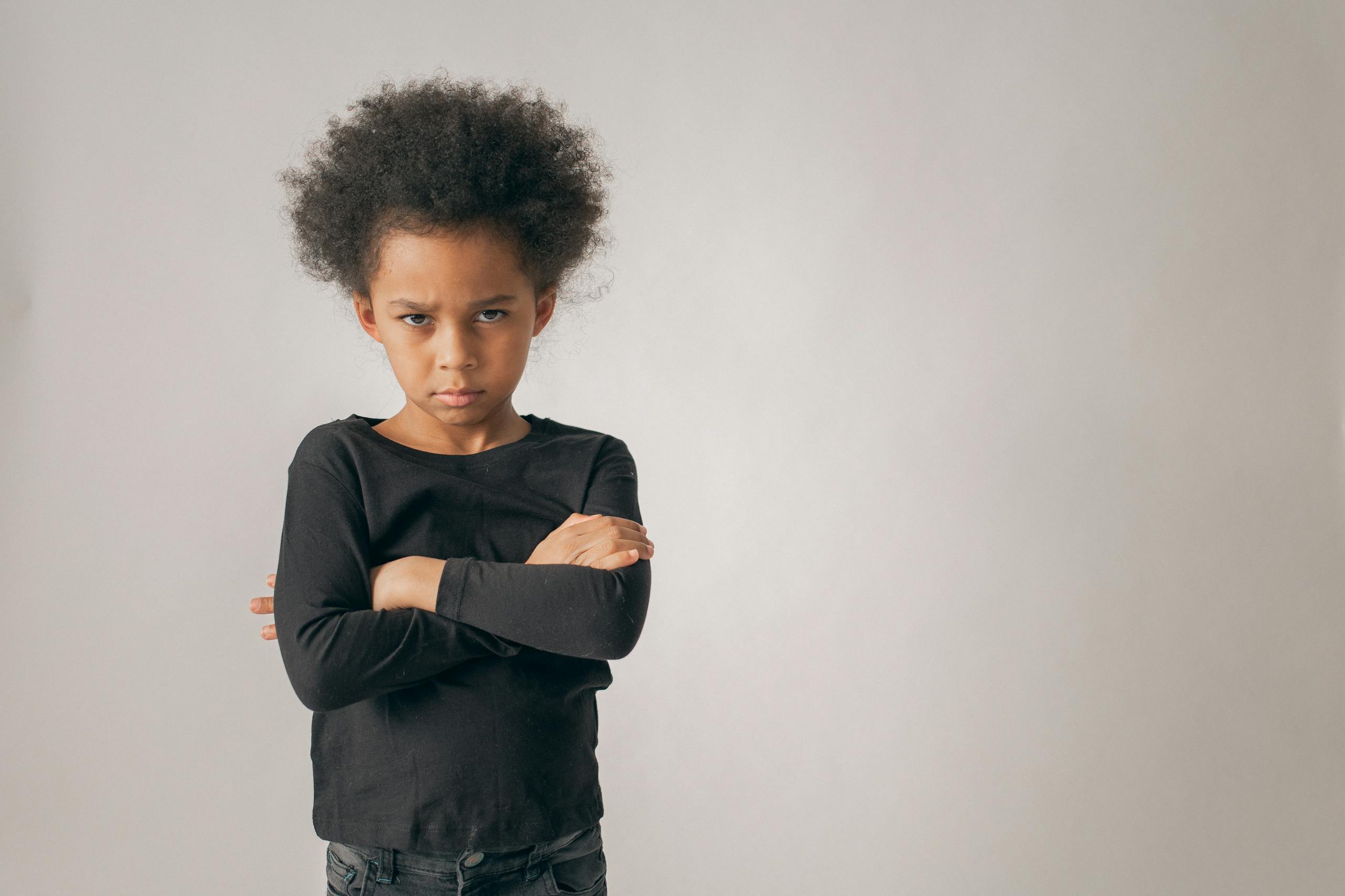 Upset African American girl with crossed arms looking at camera with serious face while standing on white background in light studio