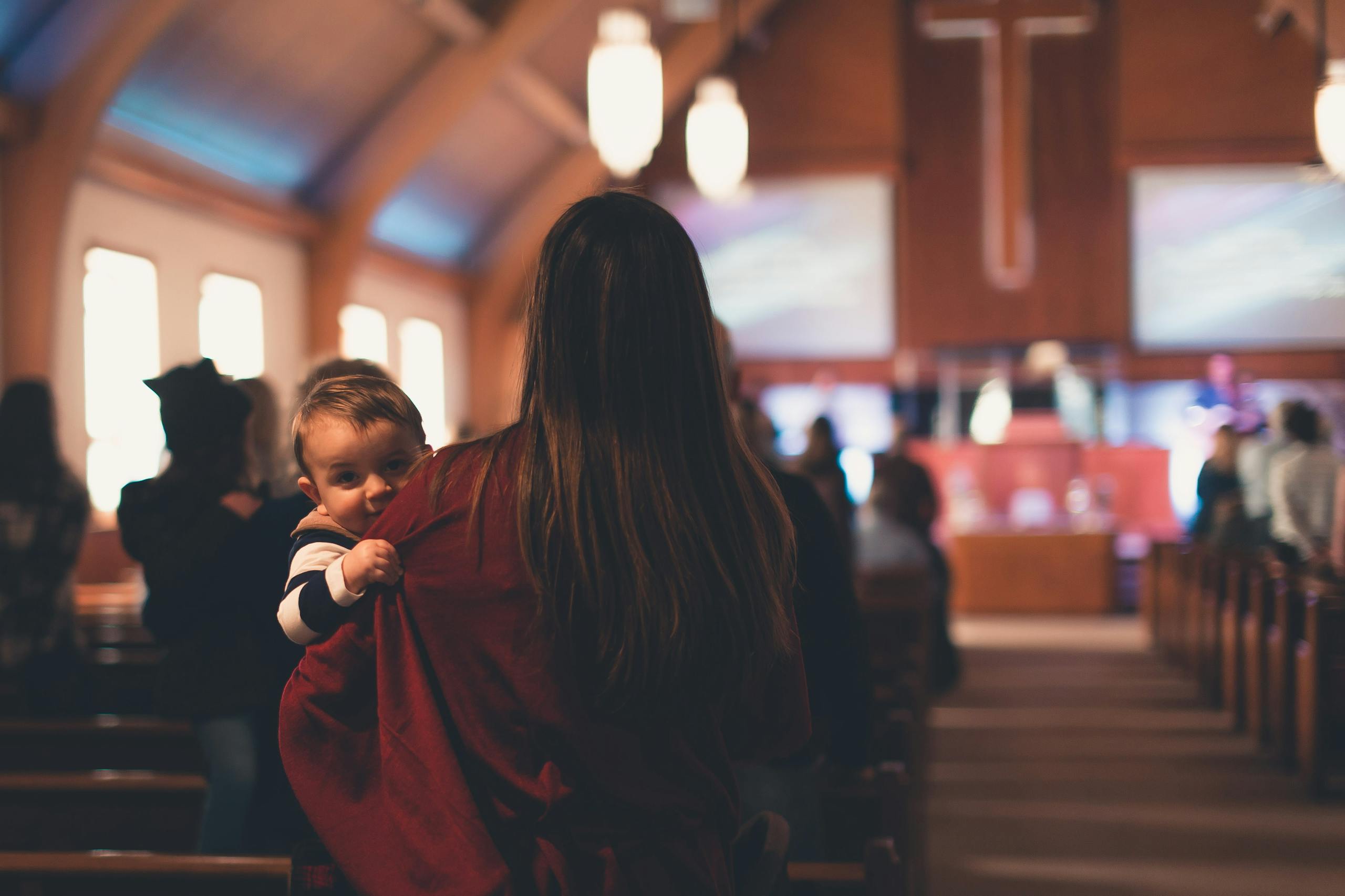 Woman carrying a child inside a church with people worshipping in the background.