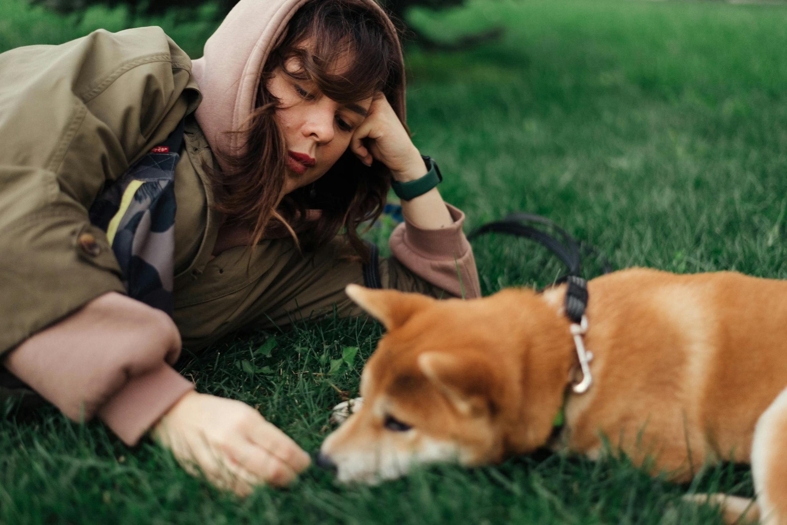 A woman in a jacket relaxing on grass with her Shiba Inu dog on a leash.