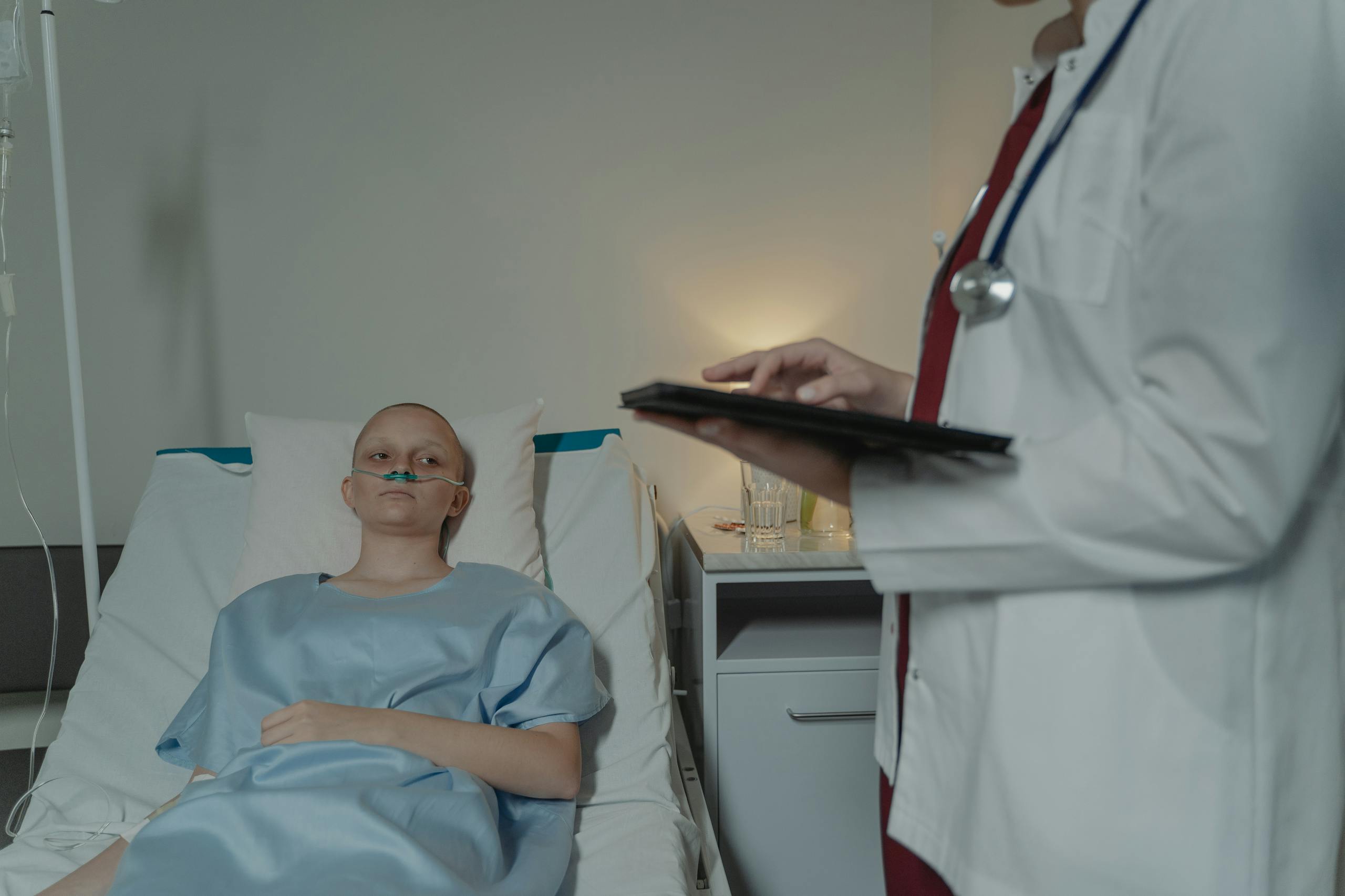 Woman in hospital gown lying in bed, doctor checks digital tablet nearby.
