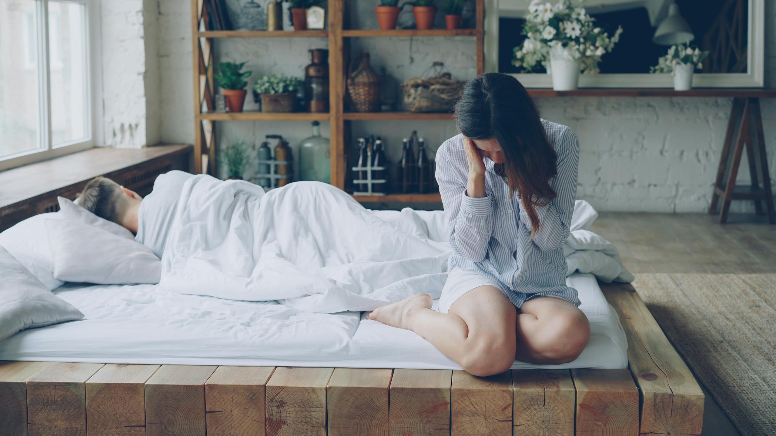 Woman in pajamas sits thoughtfully on a bed beside a sleeping person, morning light.