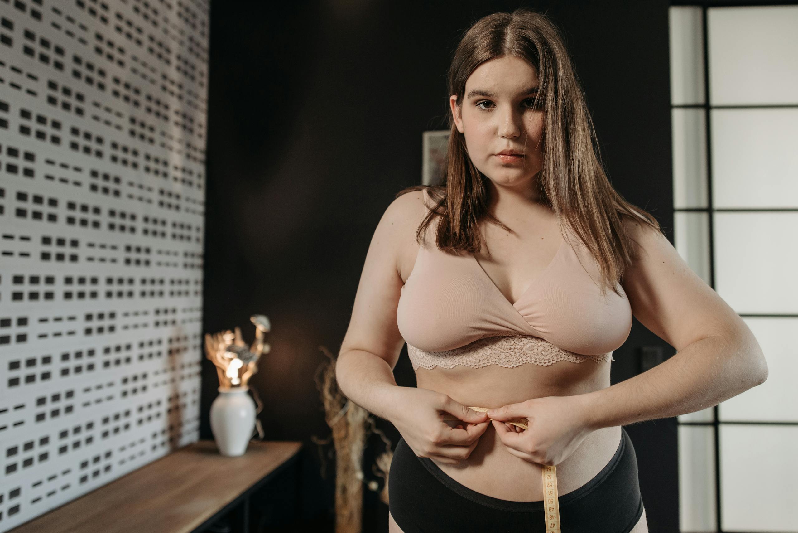 Woman in underwear measuring her waist with a tape indoors, focusing on health and self-care.