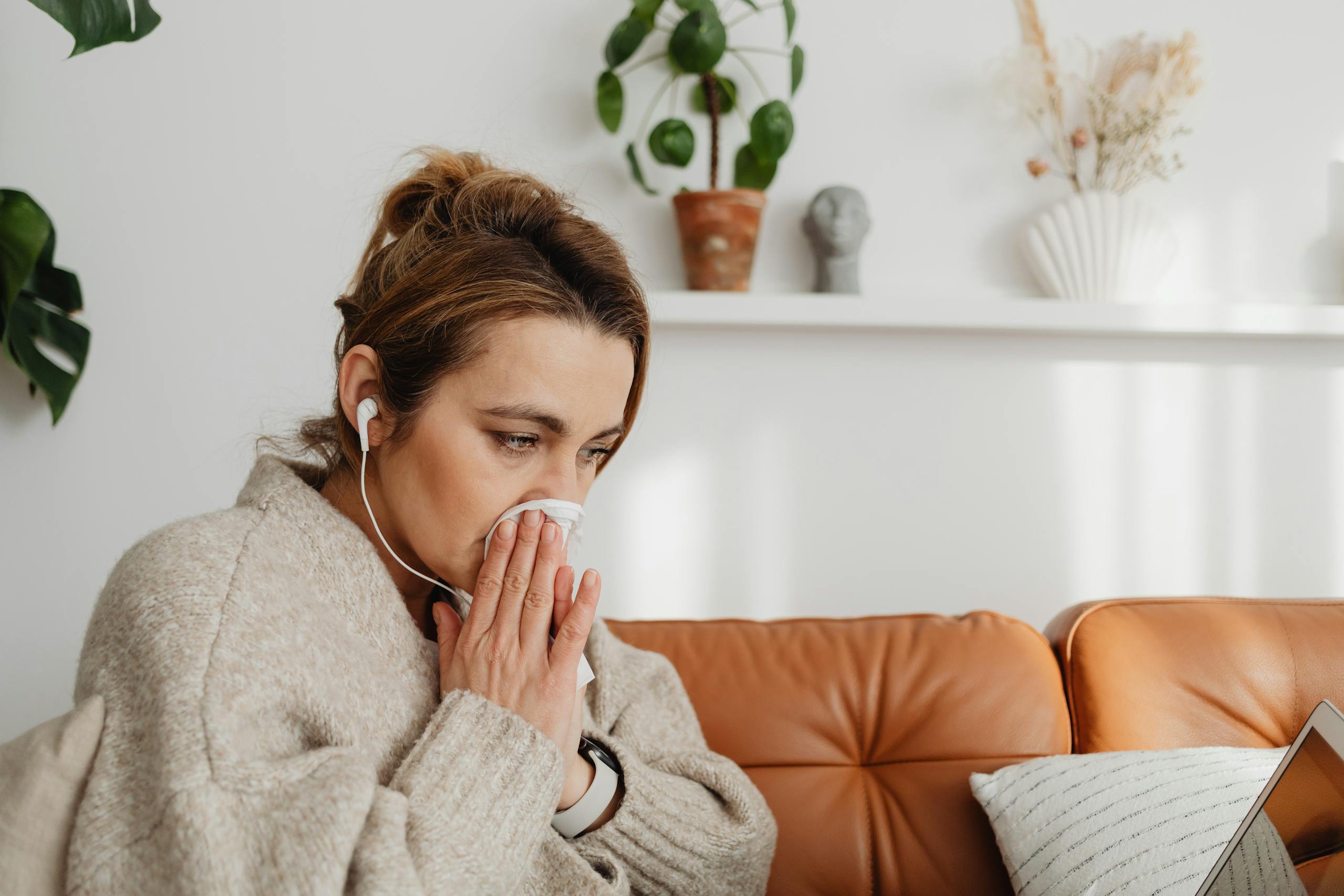 Woman sitting on a couch with earphones, holding a tissue. Indoor cozy setting.