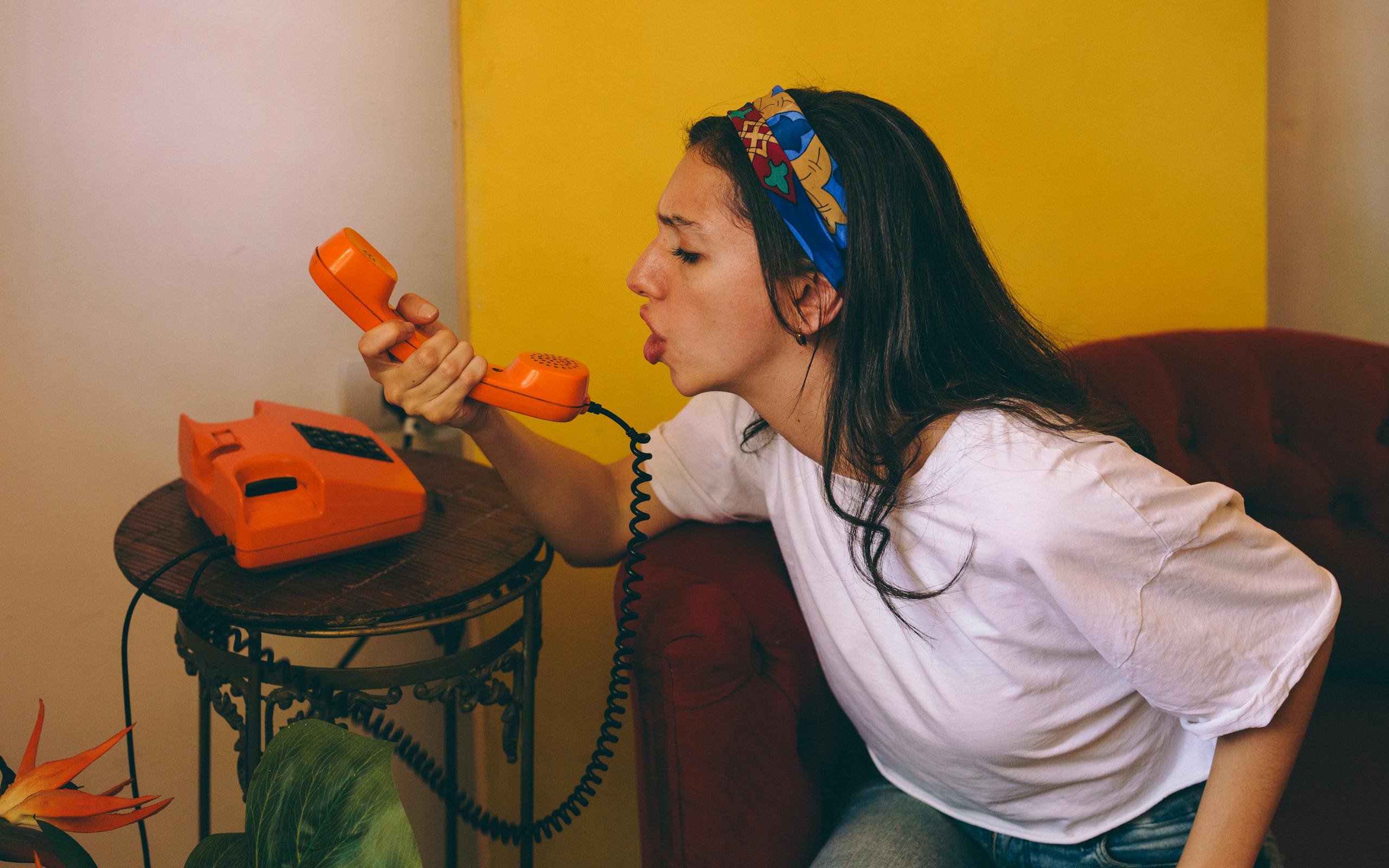 Woman speaking into a bright orange rotary phone indoors, expressing emotion.