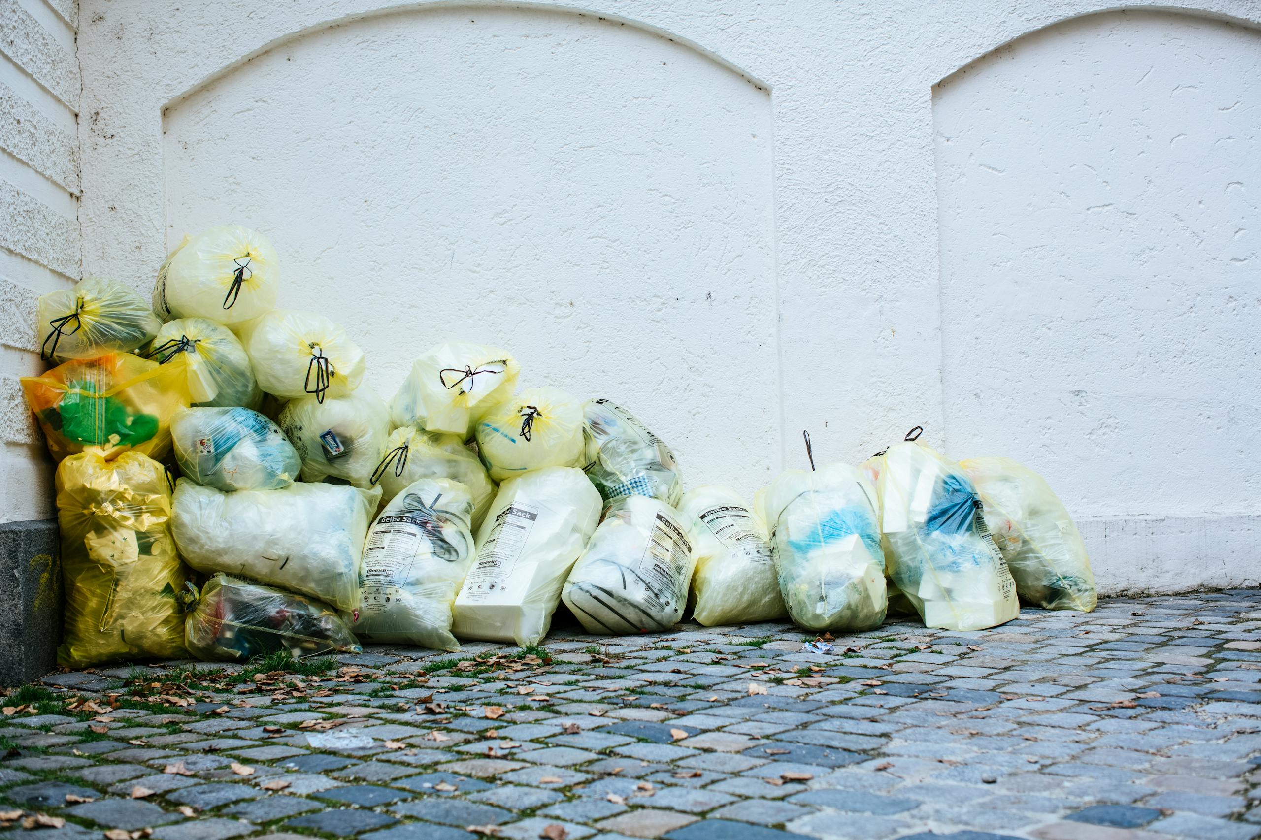 Yellow garbage bags piled against a white wall on a cobblestone street corner outdoors.