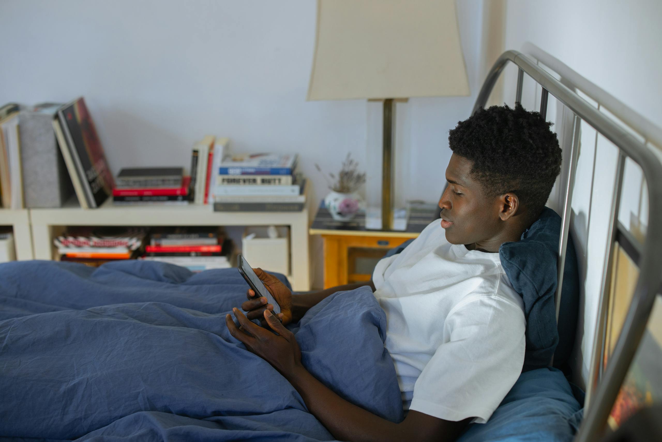 Young black man lying in bed in a modern room using his smartphone. Cozy and relaxed atmosphere.