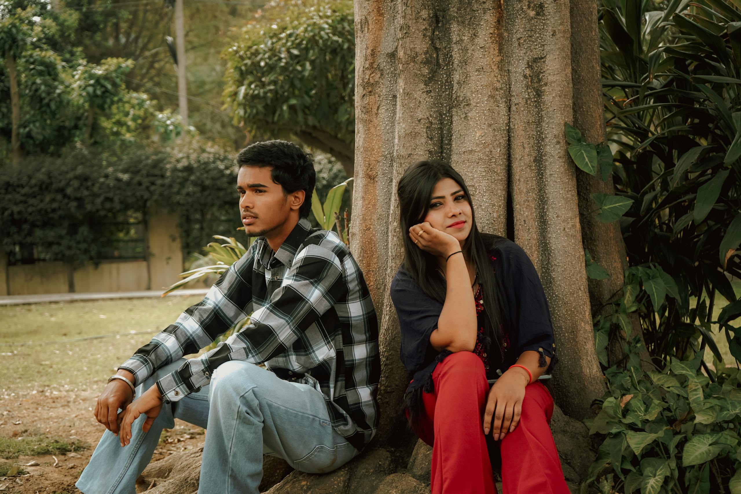 Young couple sitting pensively under a tree in a lush park setting.