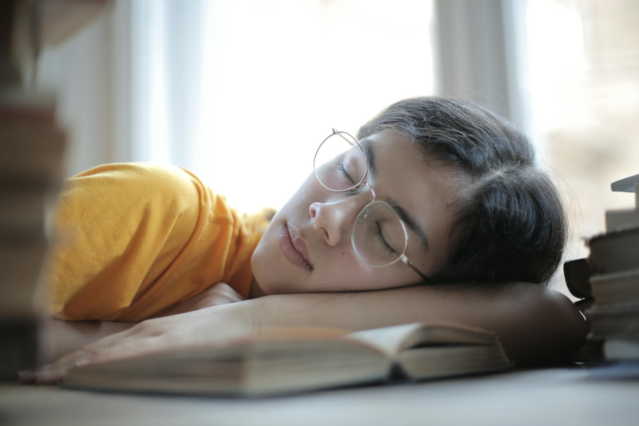 Young ethnic woman in eyeglasses and casual wear tired of exam preparation lying asleep on table with stacks of books