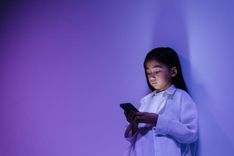 Young girl engrossed in smartphone against a vibrant purple wall indoors.
