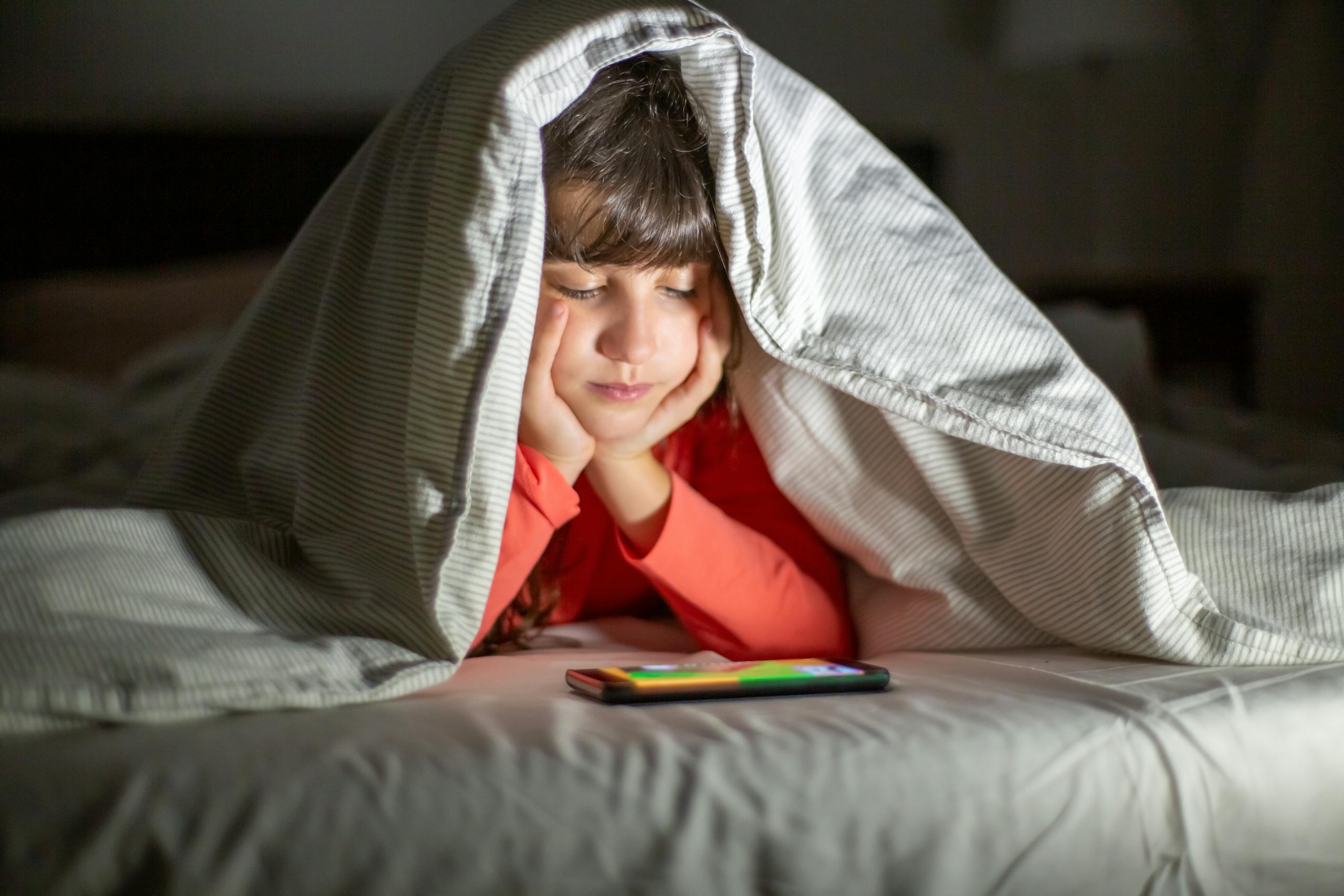 Young girl under a blanket watching a smartphone, enjoying a cozy indoor setting.