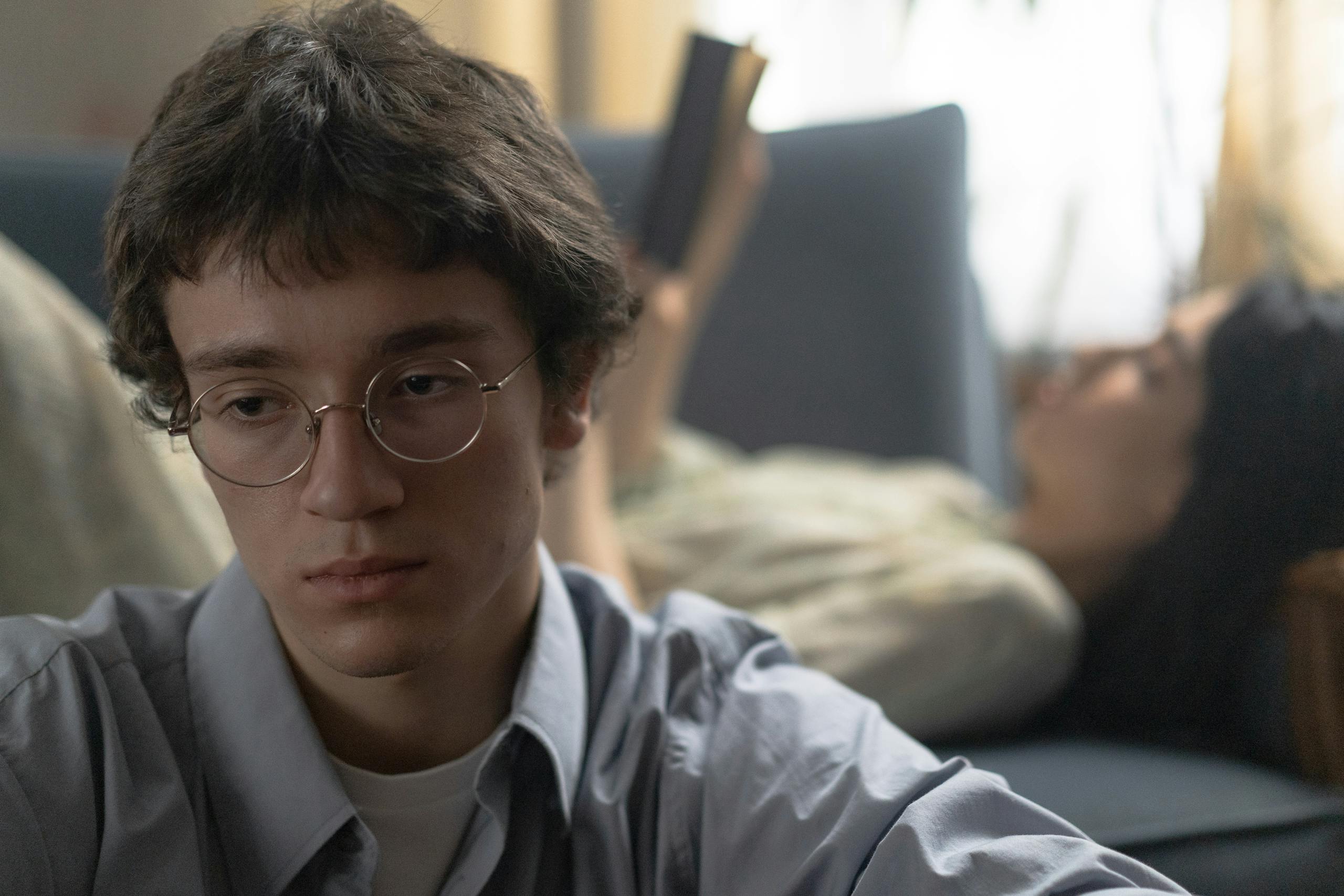 Young man in glasses looks thoughtful while a woman reads in the background, indoors.
