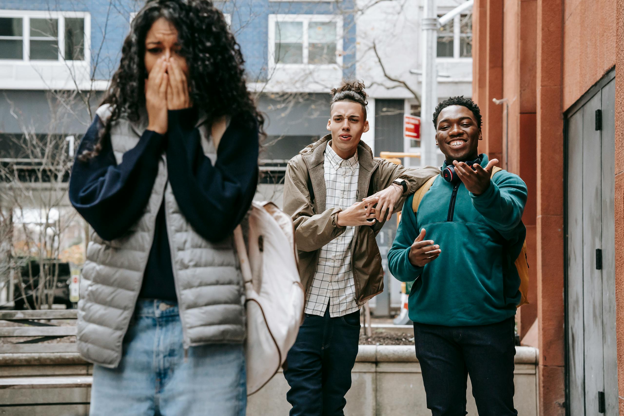 Young multiethnic male students laughing at upset female classmate covering mouth with hands and crying on street