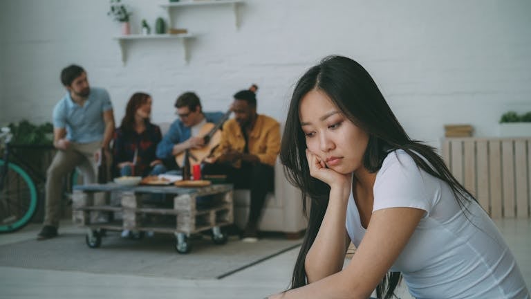 Young woman looks lonely at a party while others socialize in the background.