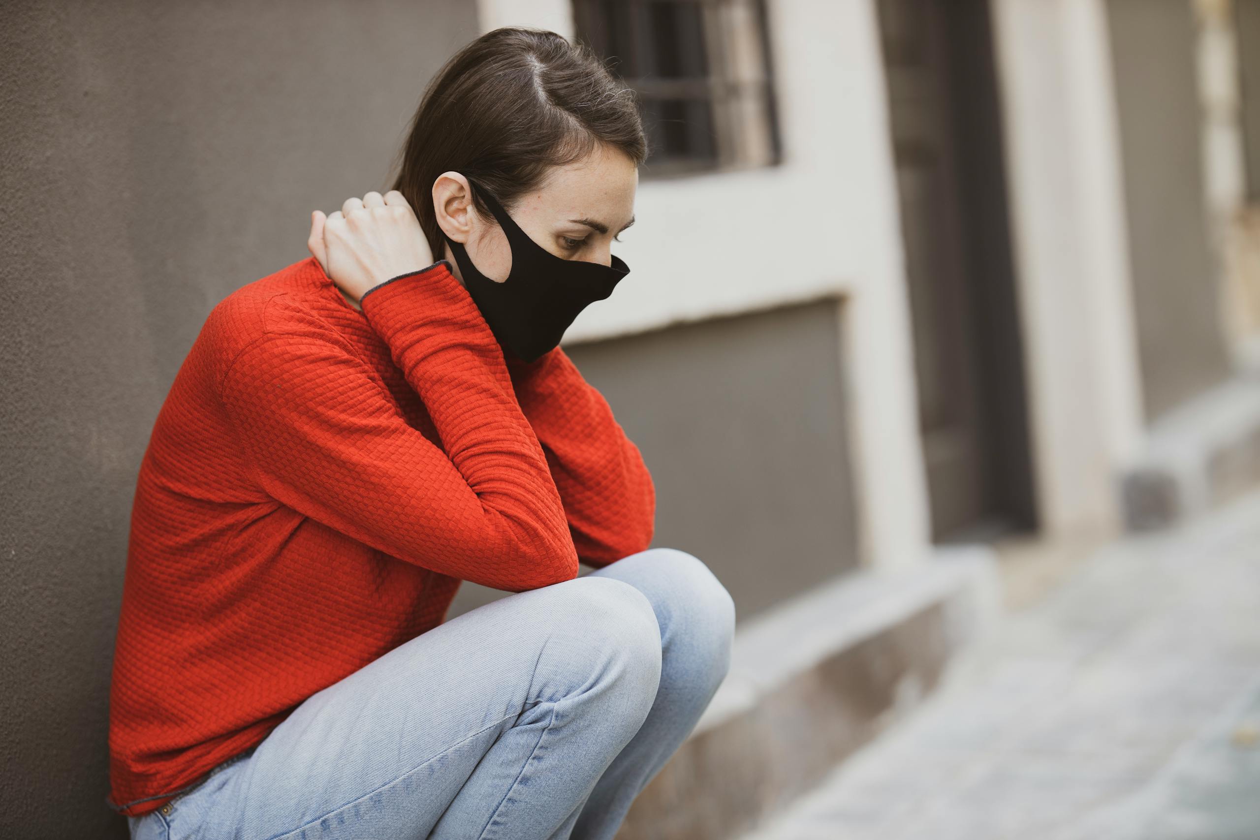Young woman wearing a face mask and red sweater sitting outdoors, symbolizing personal protection and solitude.