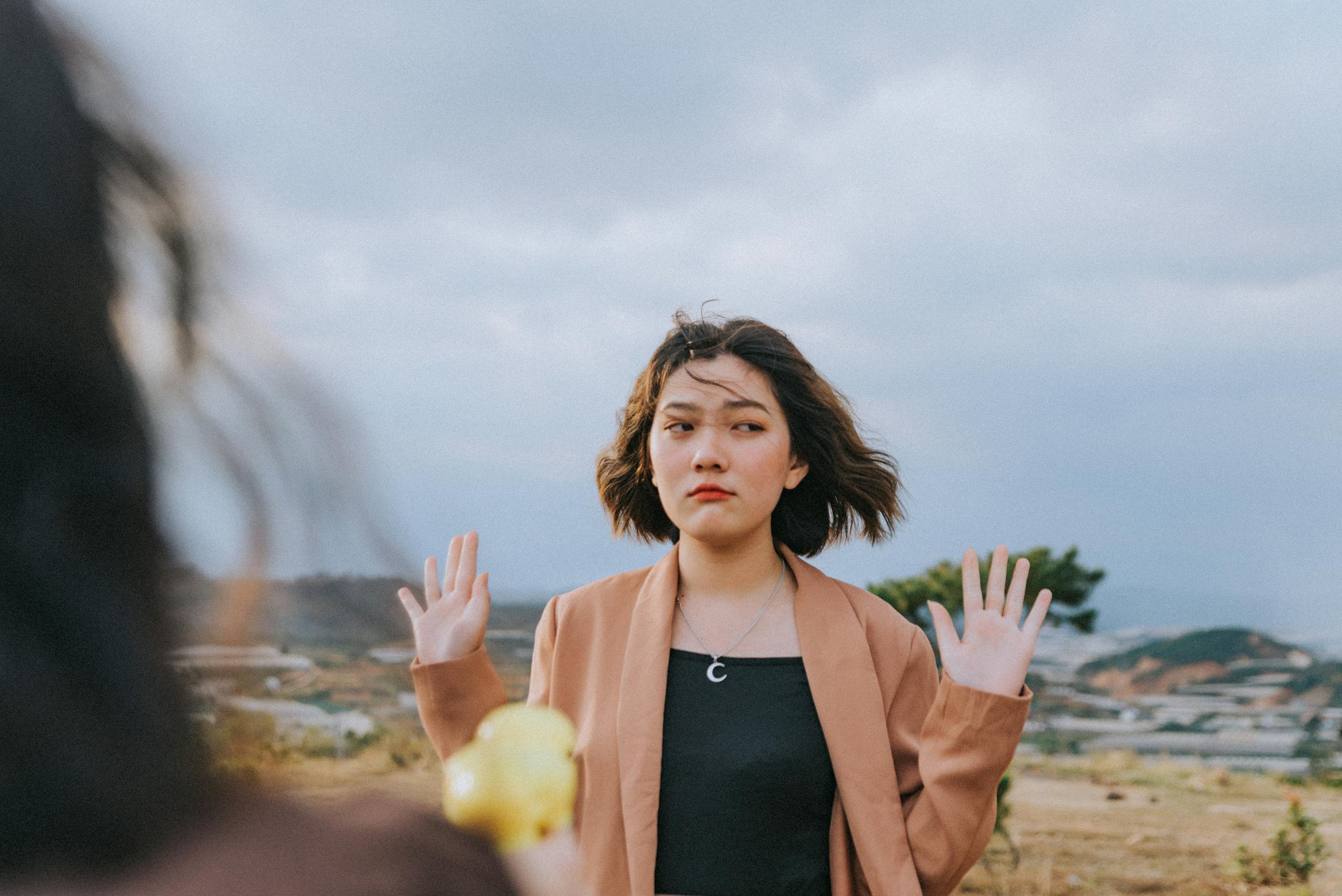 Young woman with short hair and raised hands outdoors expressing emotion. Overcast sky in background.