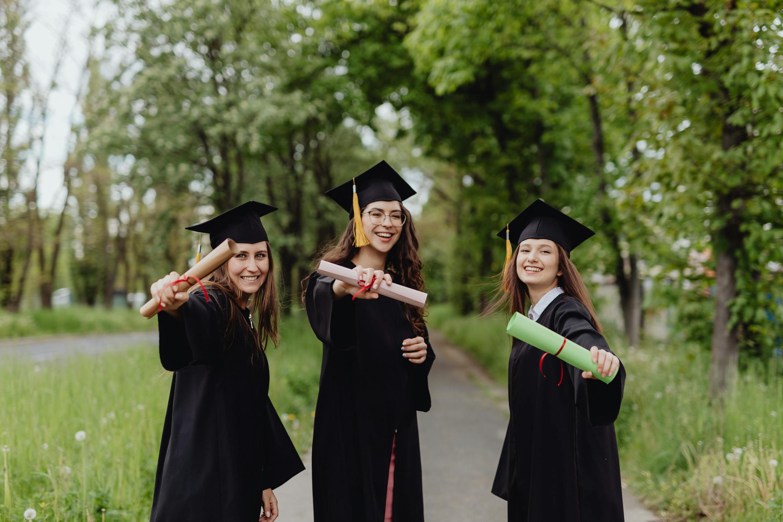 Young women in graduation gowns celebrating with diplomas in a park.