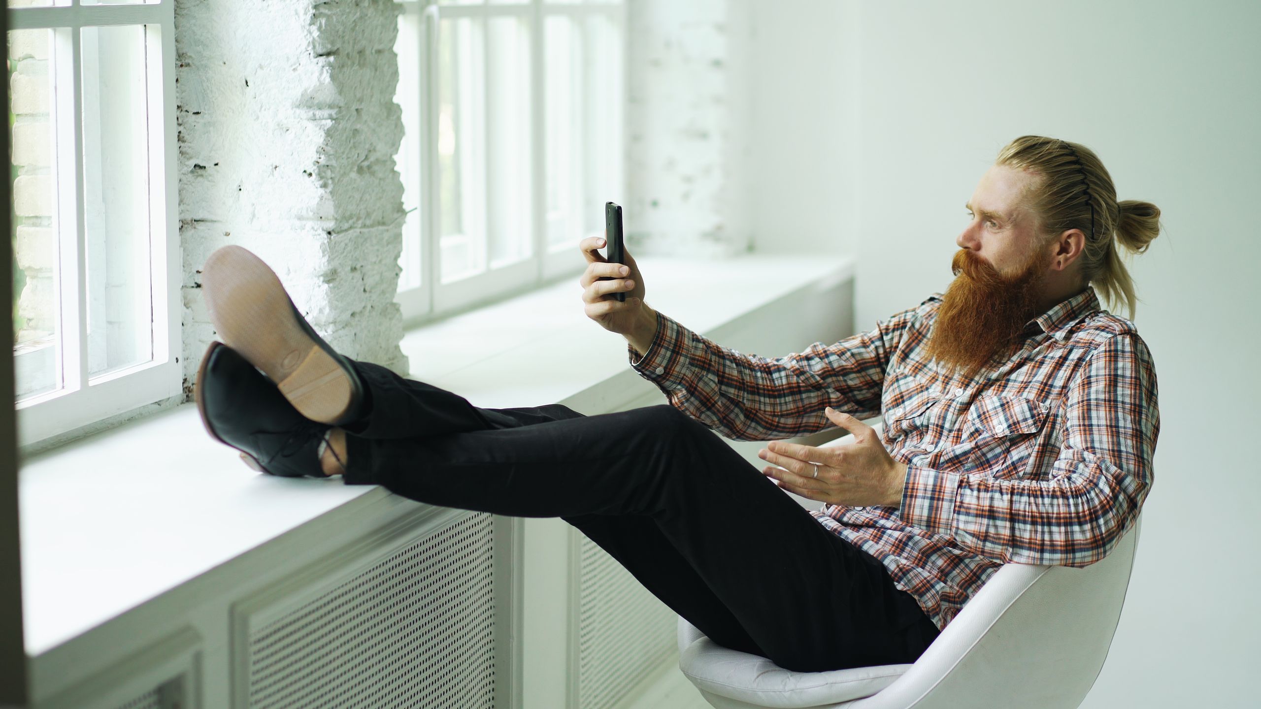 A bearded man in casual attire relaxes in a chair while making a video call indoors.