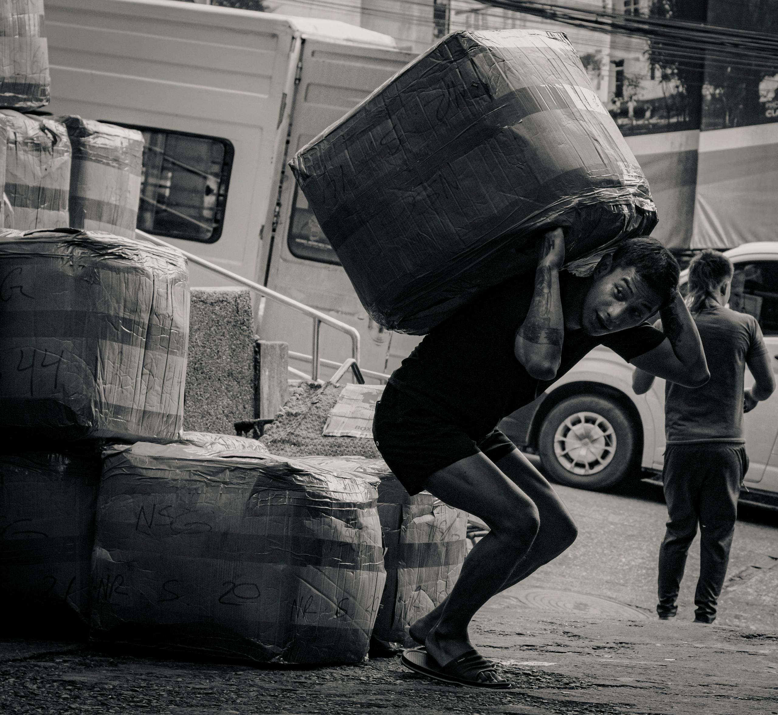 A black and white photo capturing a porter carrying a large package at a market in Baguio, Philippines.