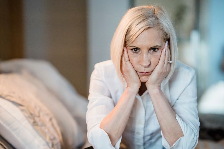 A blond woman sits thoughtfully with her hands on her chin, wearing a white blouse indoors.