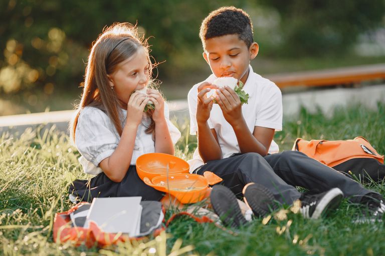 A boy and girl in school uniforms enjoying sandwiches during a picnic in the park on a sunny day.