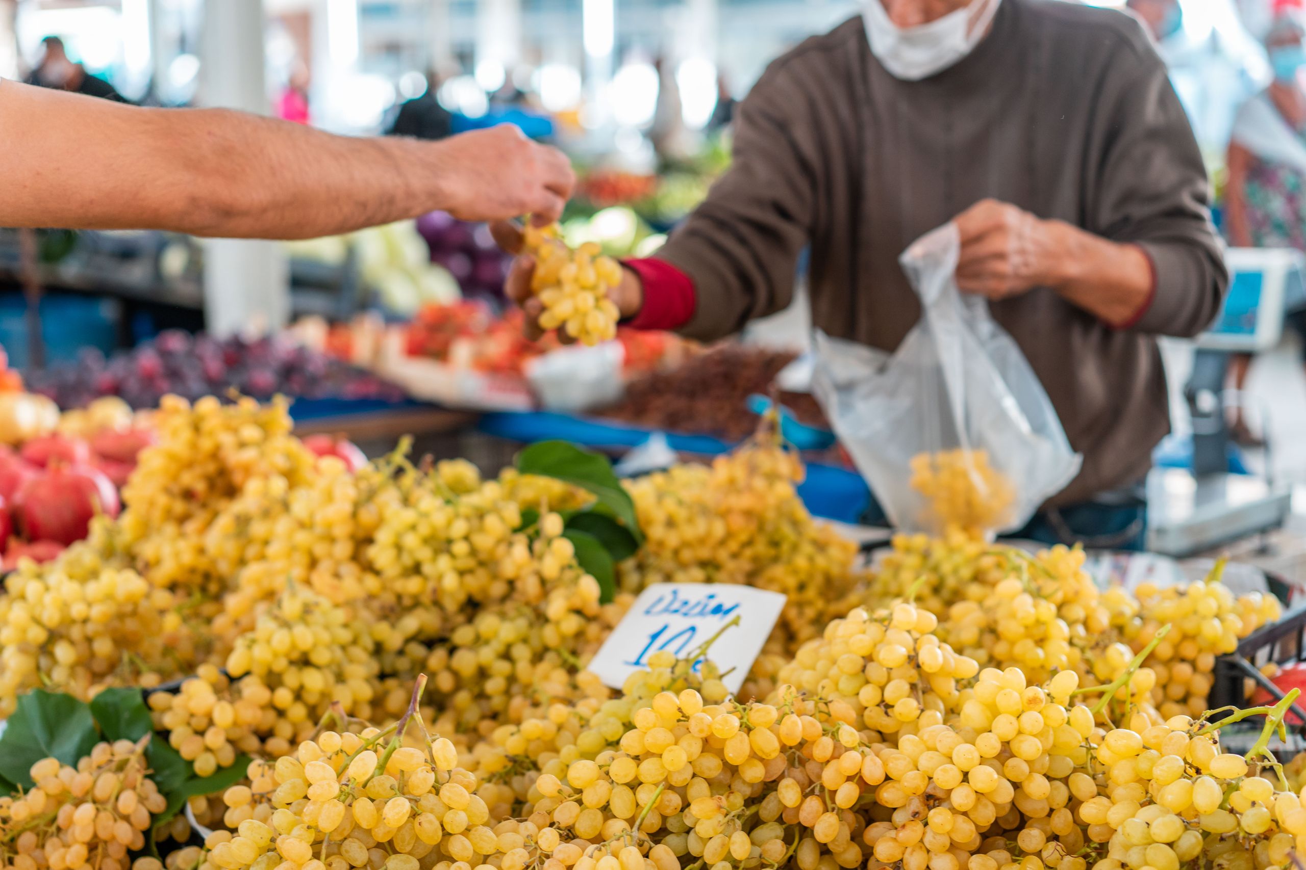 A bustling street market stall with fresh grapes being purchased. A seller hands a bunch to a buyer in a lively outdoor setting.