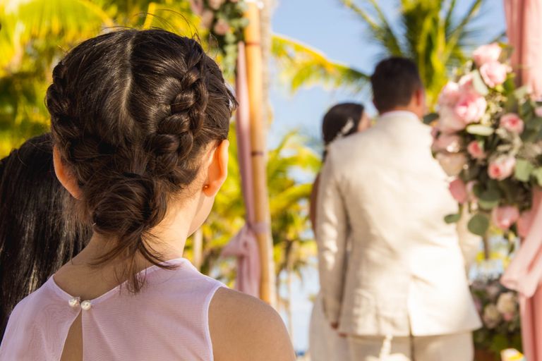 A child observes a wedding ceremony on a tropical Cancún beach with palm trees and flowers.