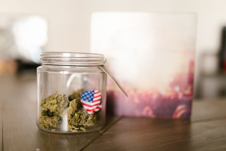 A close-up of a glass jar containing cannabis with an American flag symbol pinned to it, indoors.