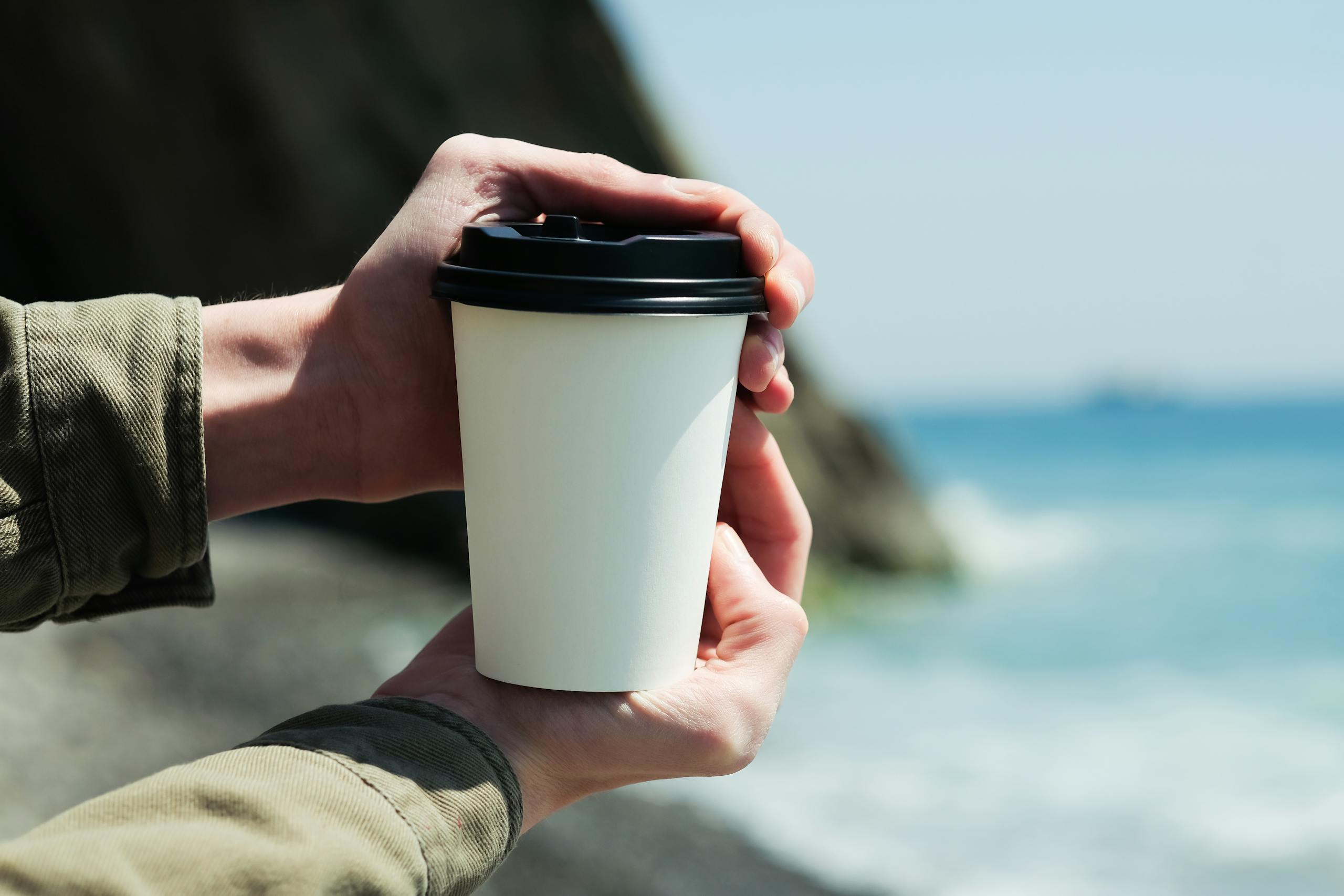 A close-up of hands holding a recyclable paper cup with a lid by the seaside.