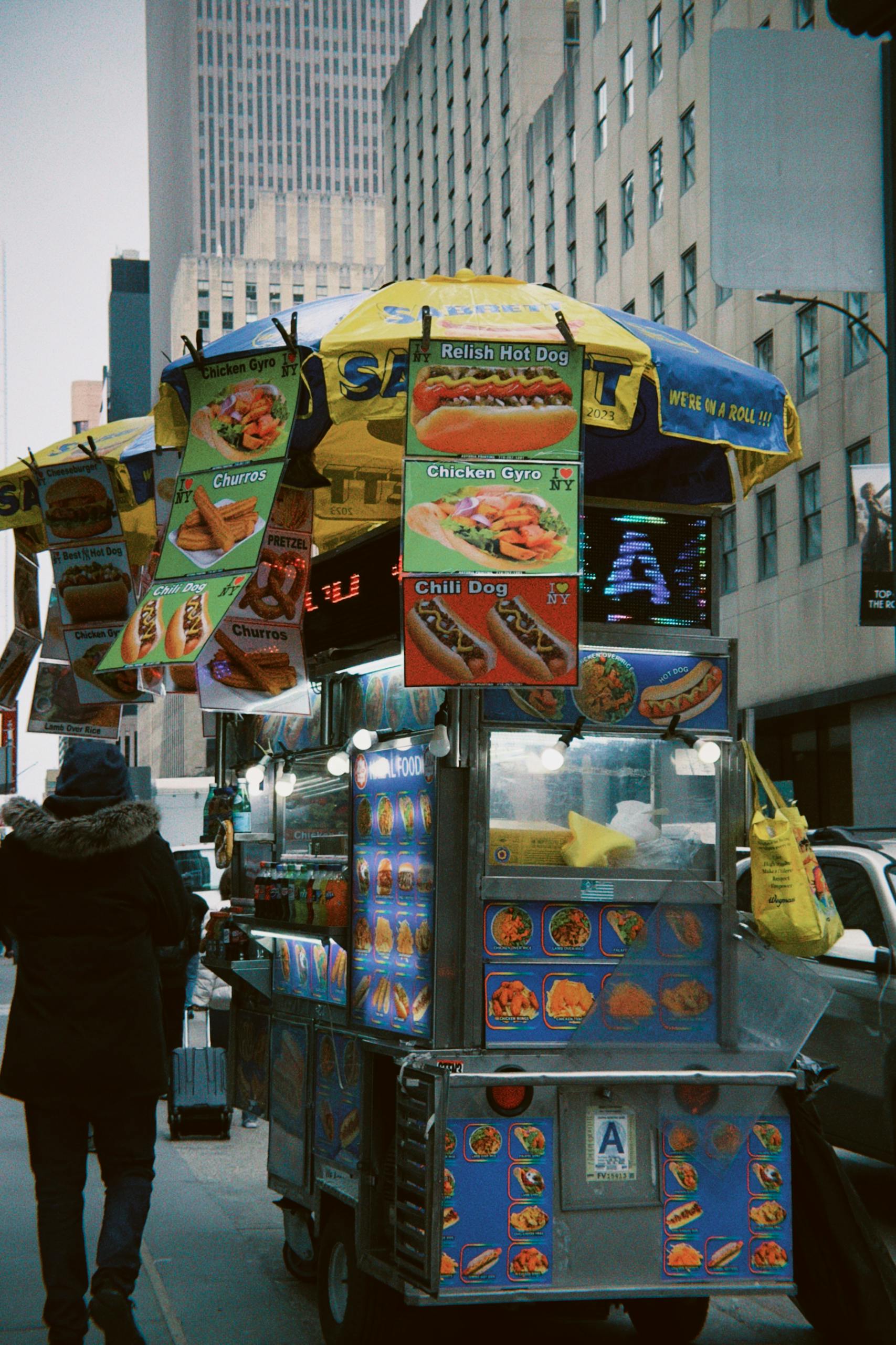 A colorful food cart offers hot dogs, churros, and more on a bustling NYC street.