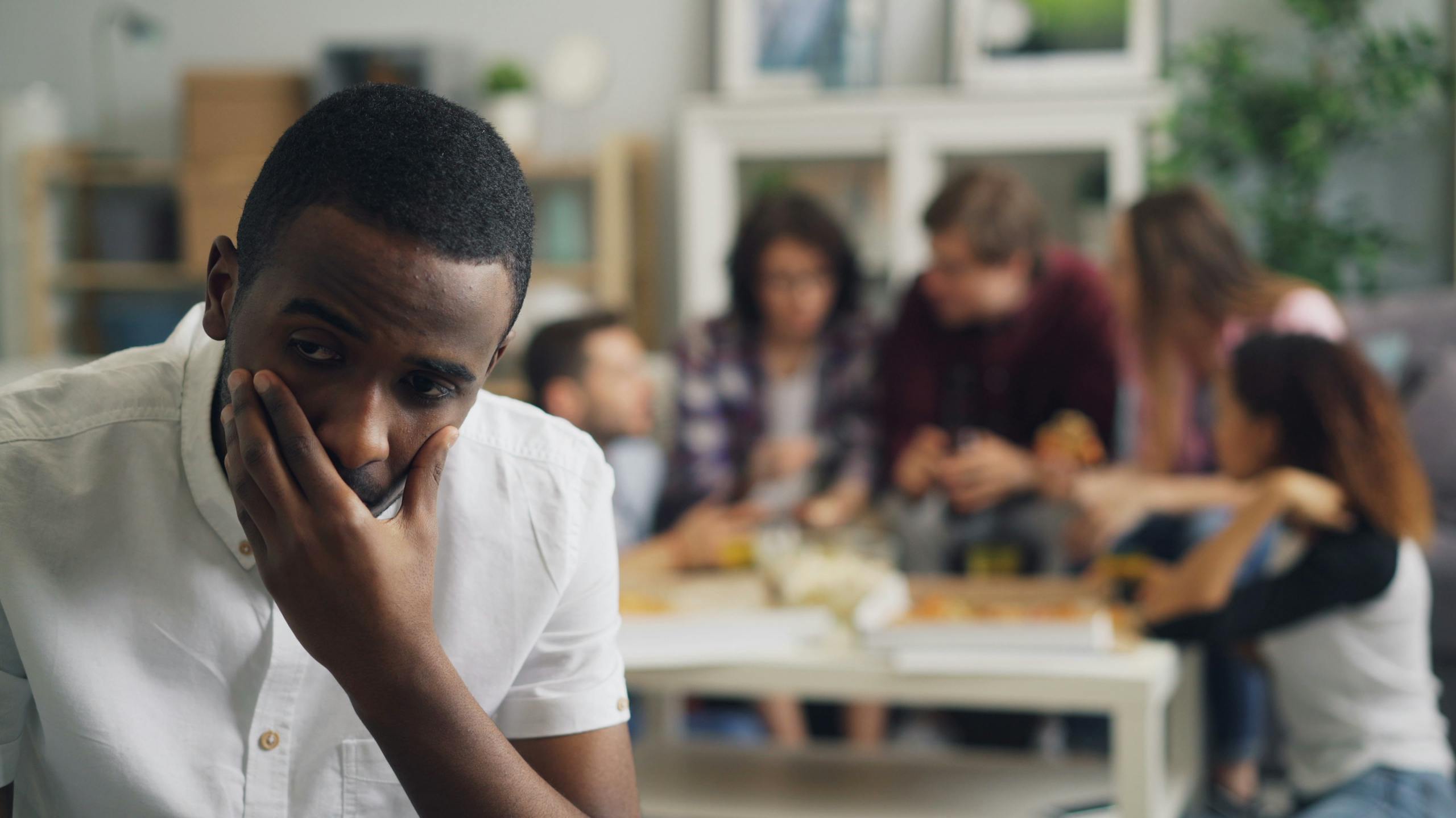 A concerned man sits apart from a lively group in a cozy home setting.