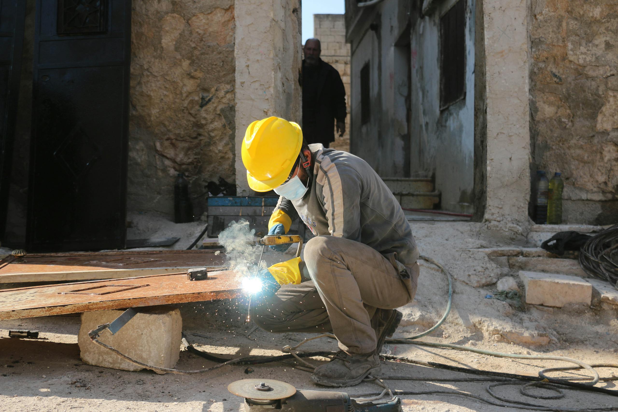 A construction worker welding outdoors in Idlib, Syria, wearing safety gear and a hard hat for protection.