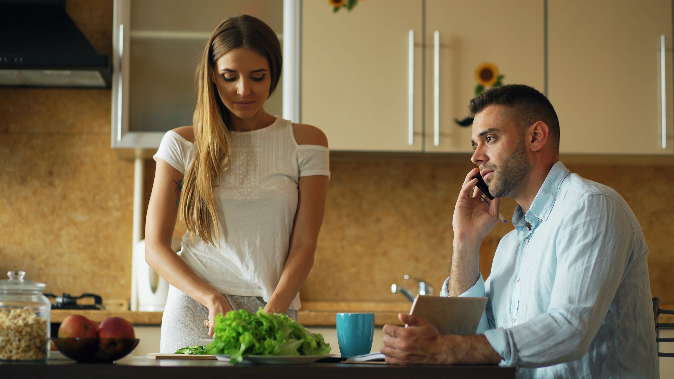 A couple in a kitchen, one preparing salad, the other on the phone, showcasing lifestyle balance.