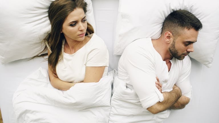 A couple in bed, facing away with tense expressions, suggesting conflict.