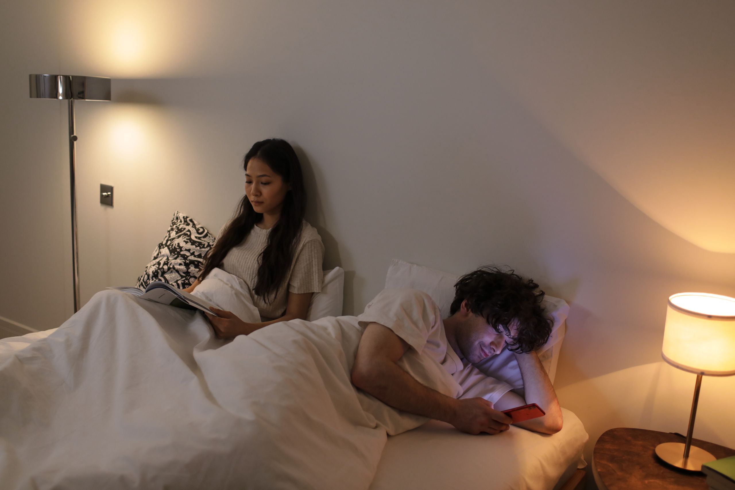 A couple in bed, one reading and the other using a phone, with warm lighting.