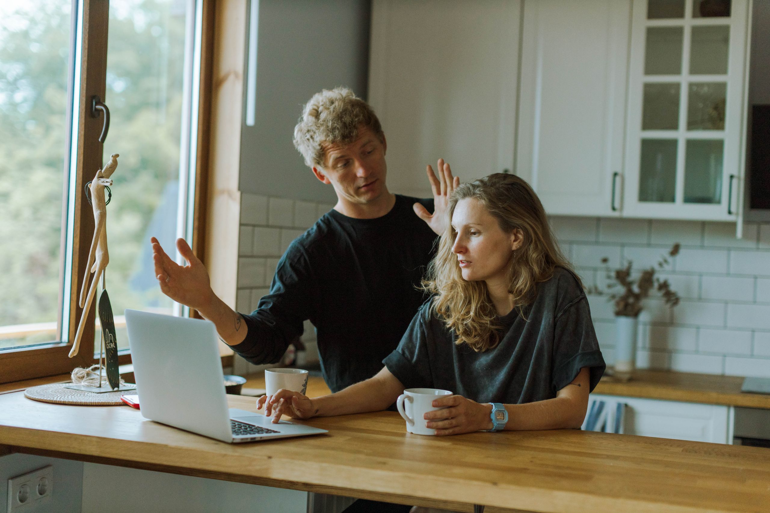 A couple interacting in a bright kitchen using a laptop at a wooden counter.