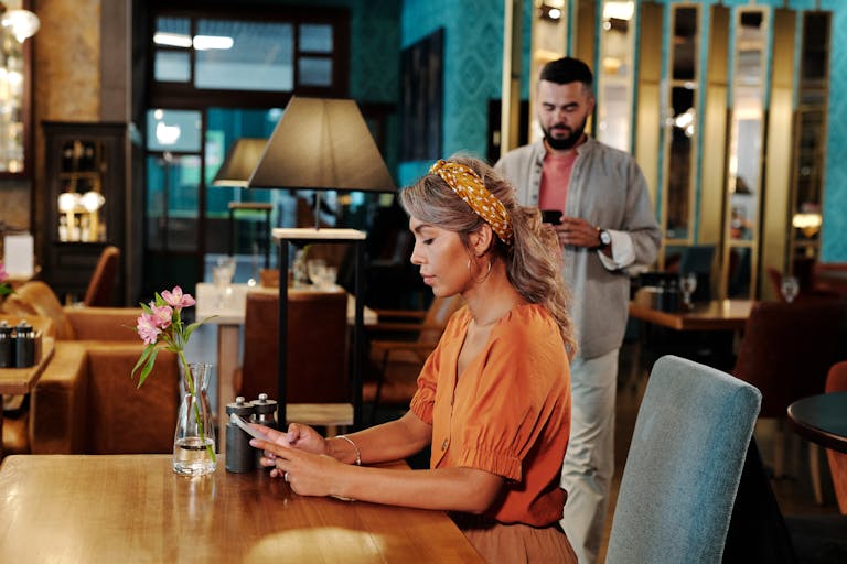 A couple on a first date at a modern café. The woman awaits, using her smartphone.