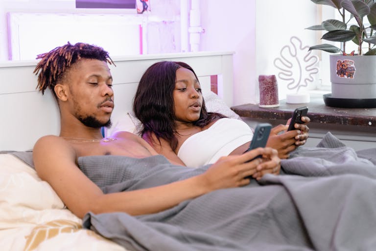 A couple relaxes in bed, focused on their phones, in a cozy indoor setting.
