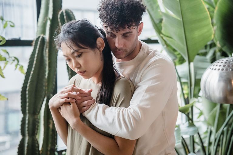 A couple shares an intimate back hug surrounded by large indoor plants.