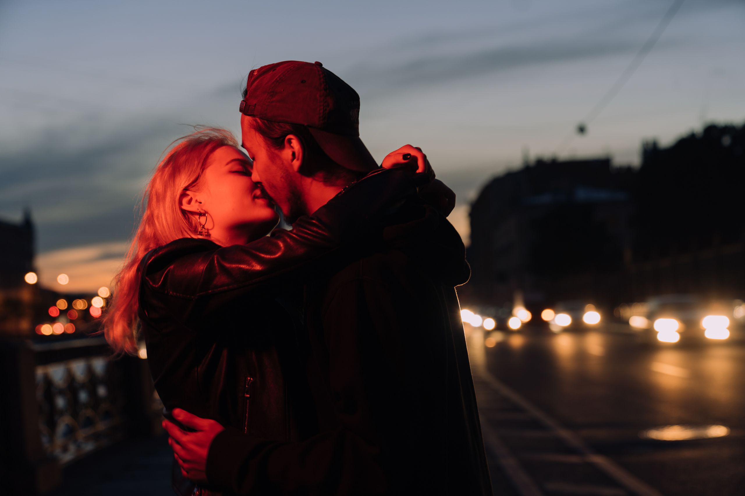 A couple sharing an intimate kiss on a city street during twilight, with blurred lights in the background.