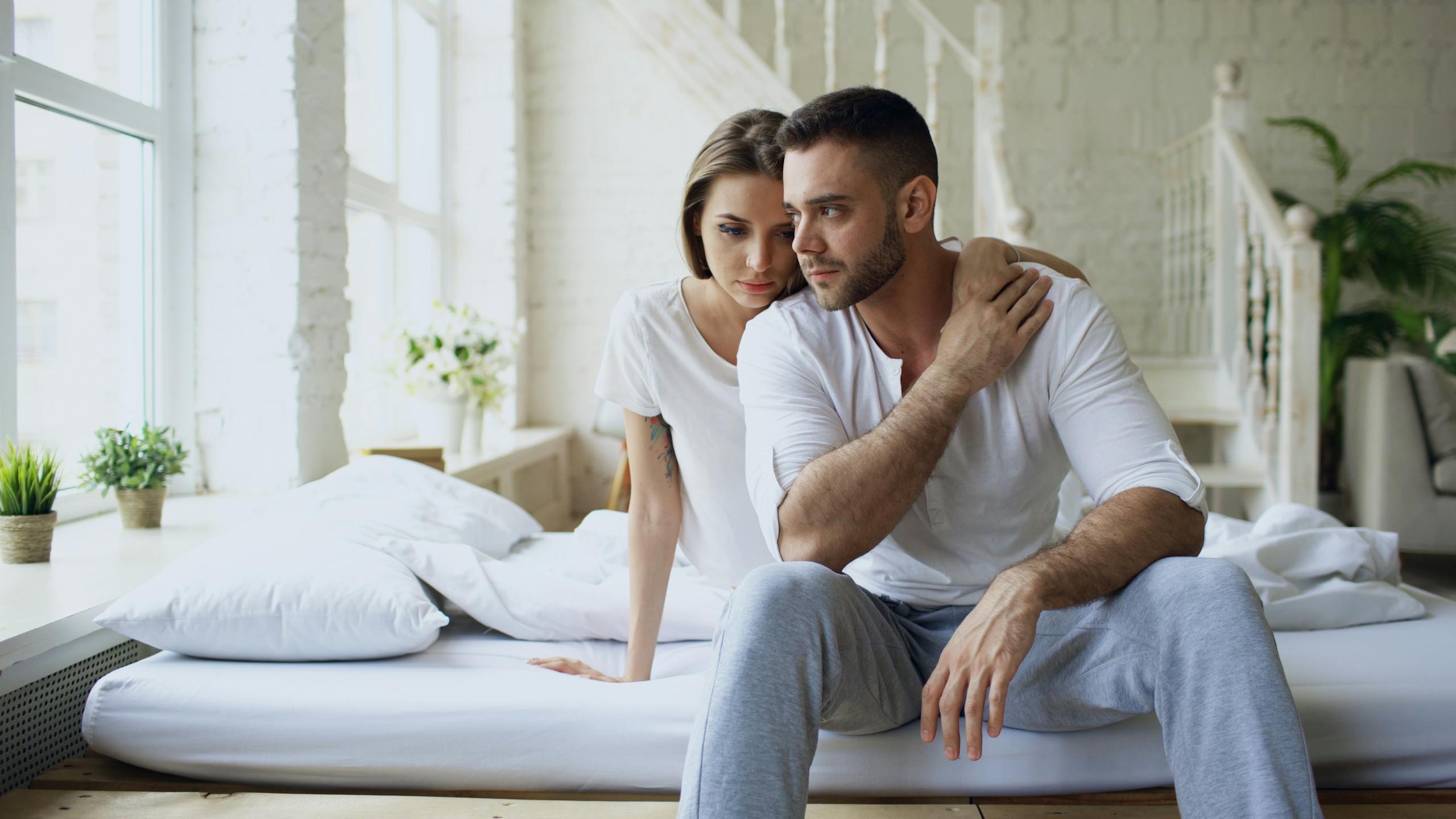 A couple sits closely together in a bright and stylish bedroom, looking serene.