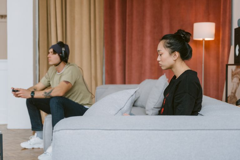 A couple sits on a couch, illustrating tension as one plays video games and the other appears distant.