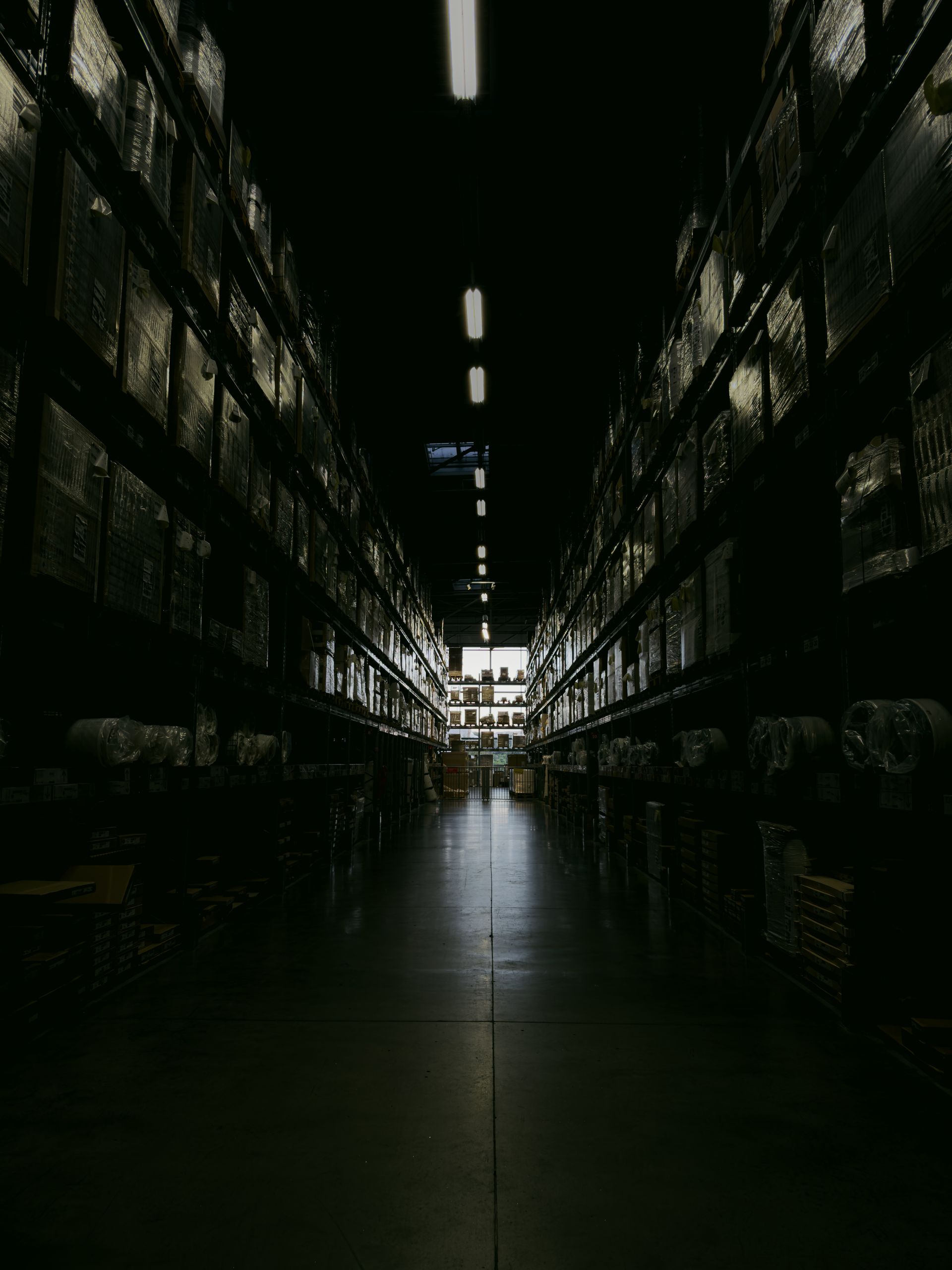 A dark, narrow warehouse aisle with shelves filled with boxes and goods, illuminated by overhead lights.