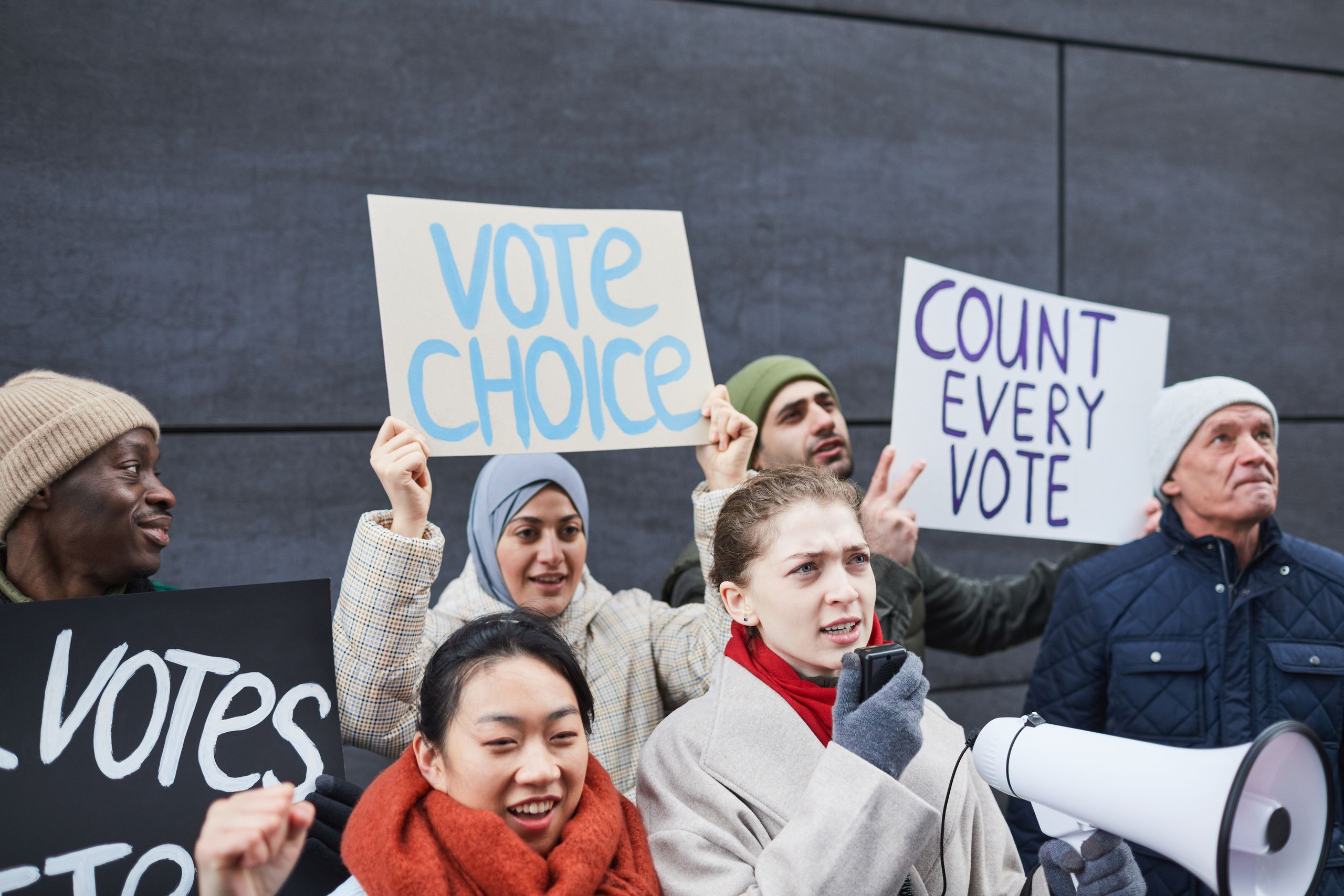 A diverse group of adults hold signs demanding vote counts and choice at an outdoor protest.