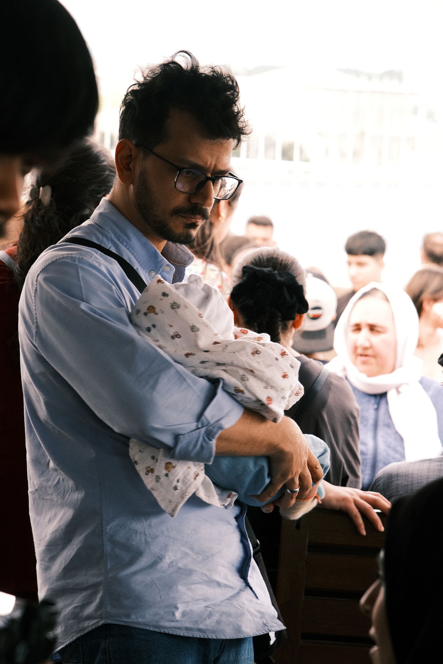 A father holds his baby protectively amidst a diverse crowd outdoors during the day.