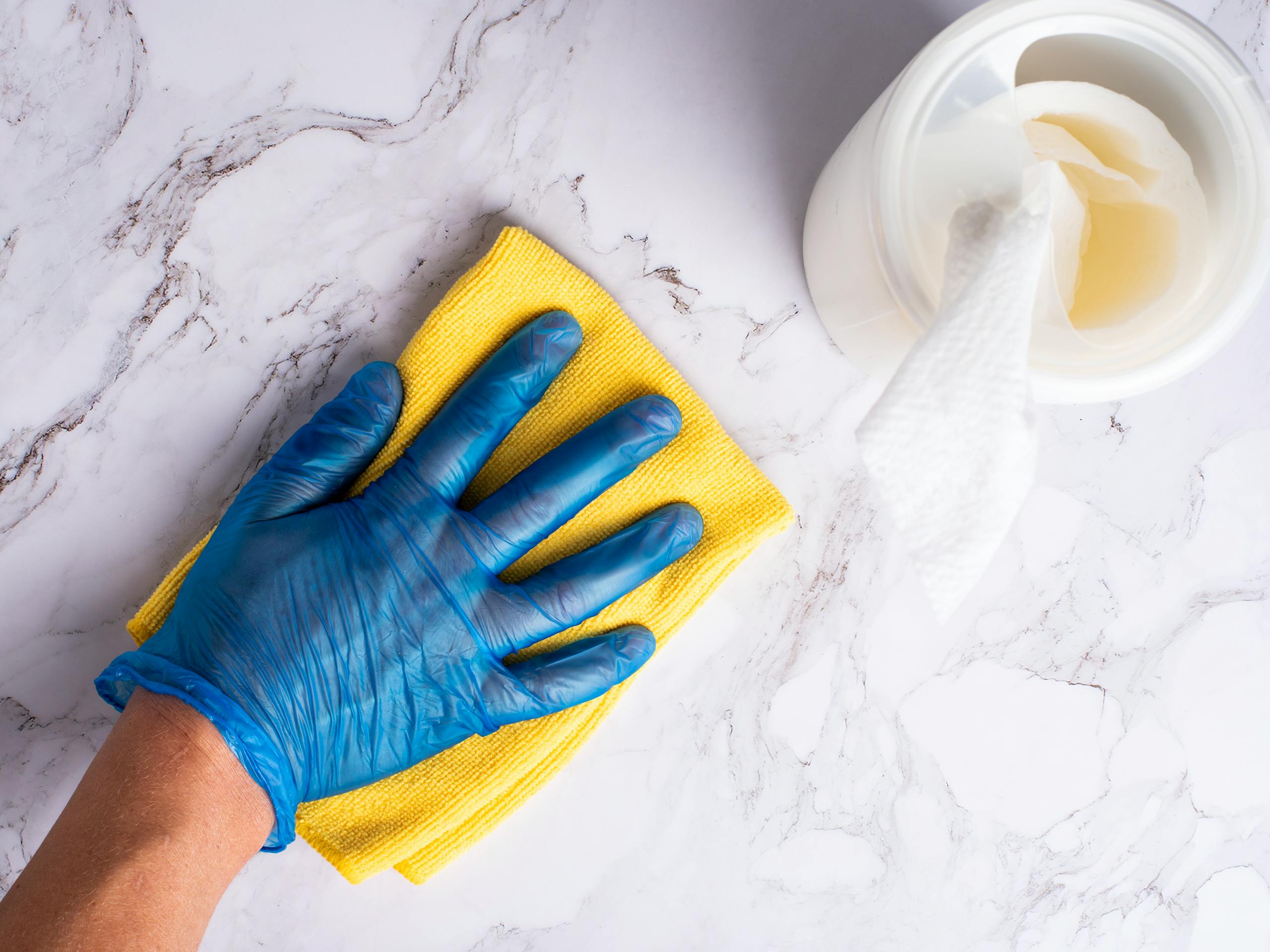 A gloved hand cleans a marble countertop with a yellow cloth next to disinfectant wipes.