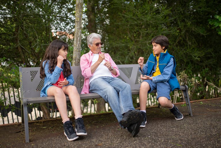 A grandfather sits with his grandchildren on a park bench, surrounded by nature, enjoying a sunny day.