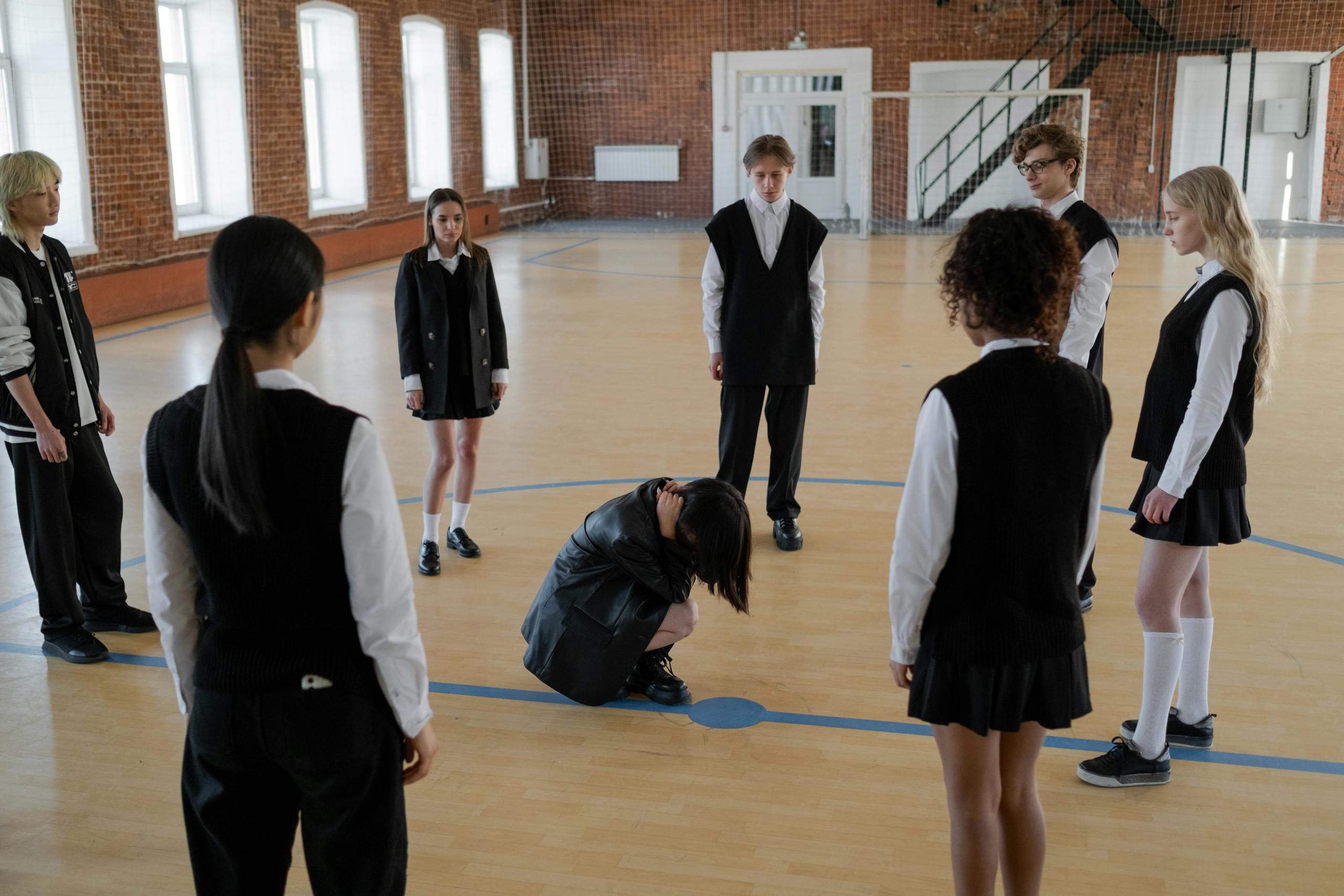 A group of teenagers in school uniforms gather around a peer in a gym setting, depicting peer dynamics.